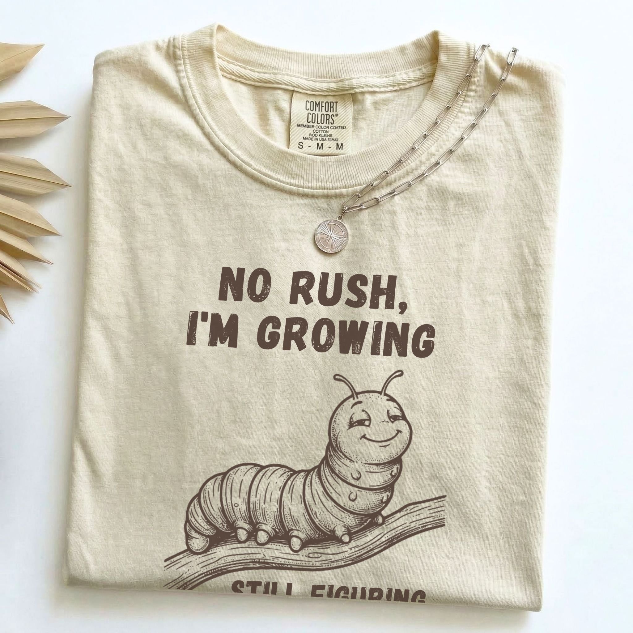 No Rush I'm Growing caterpillar Comfort Colors tee in Ivory, folded flat lay with dried palm leaves