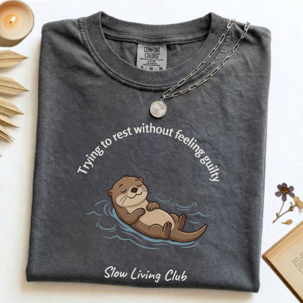 Trying to Rest Without Feeling Guilty otter 
graphic tee in Pepper – Comfort Colors unisex 
shirt flat lay – Slow Living Club collection
