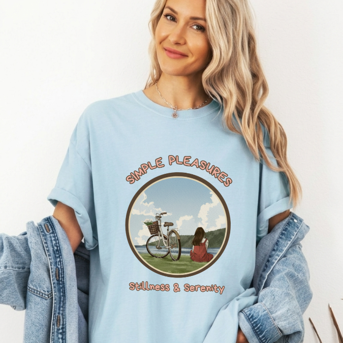 Light blue Comfort Colors graphic tee with a circular lakeside scene featuring a bicycle, a seated woman, and the phrase “Simple Pleasures Stillness & Serenity.”