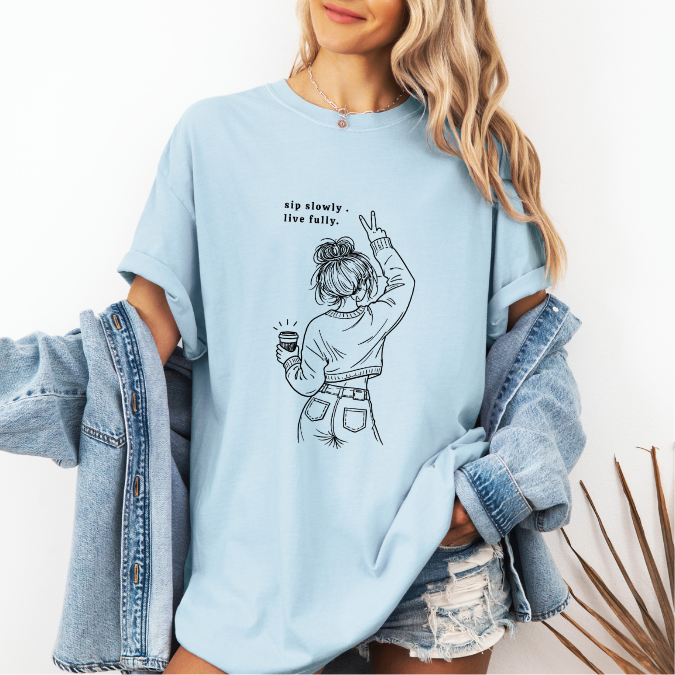 Light blue Comfort Colors graphic tee with a feminine line art illustration of a woman holding coffee and the phrase “sip slowly. live fully.”