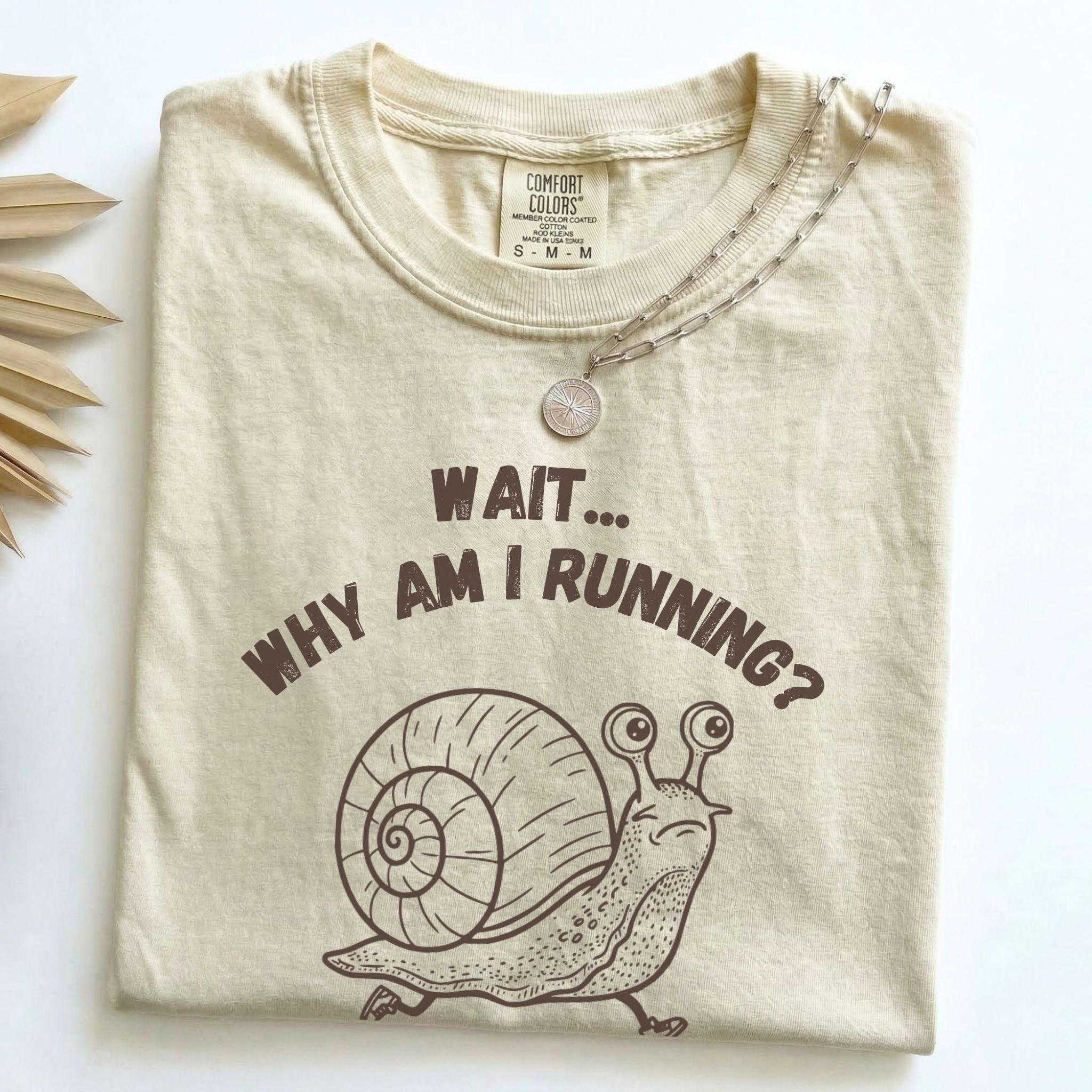 Wait Why Am I Running snail Comfort Colors tee in Ivory, folded flat lay with dried palm leaves